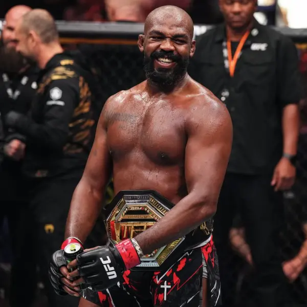 Un combattant d'arts martiaux mixtes torse nu sourit dans un octogone de l'UFC, tenant une ceinture de champion. Portant des gants noirs et un short rouge, il symbolise la victoire et la fierté.