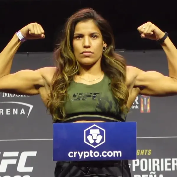 Une femme vêtue d'un haut court griffé UFC contracte ses muscles avec assurance sur scène, debout derrière un podium orné du logo Crypto.com. Expression déterminée.