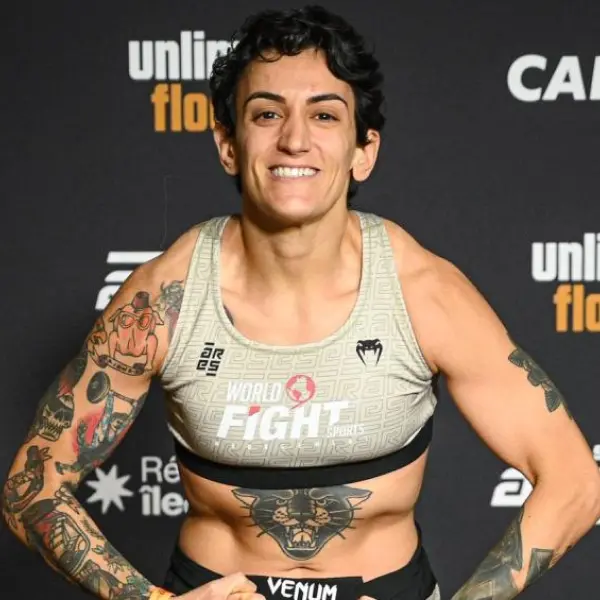 L'athlète contracte ses muscles et sourit avec assurance. Il porte un haut de sport gris avec l'inscription « World Fight Sports » et des tatouages sur les bras. Il affiche une humeur énergique et victorieuse.