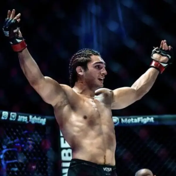 Un combattant de MMA en short noir et cheveux tressés, les bras levés par procuration dans une cage. La scène évoque le triomphe et l'adrénaline dans une arène faiblement éclairée.