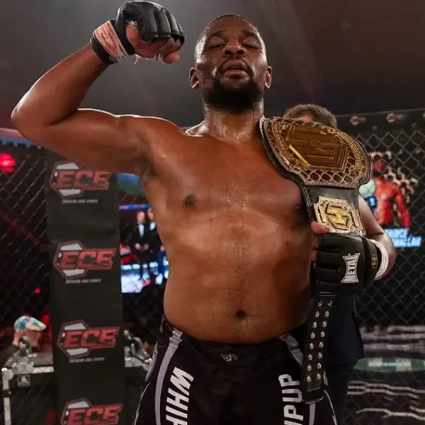 Combattant de MMA dans une cage, tenant une ceinture de champion sur son épaule. Il lève son autre poing en signe de victoire, affichant une expression déterminée et triomphante.