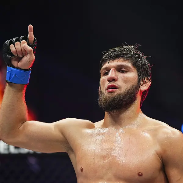 Un combattant de MMA barbu, en sueur et intense, pointe vers le ciel, signe de victoire. Sa main gantée de bleu est levée, sur fond d'arène faiblement éclairée.